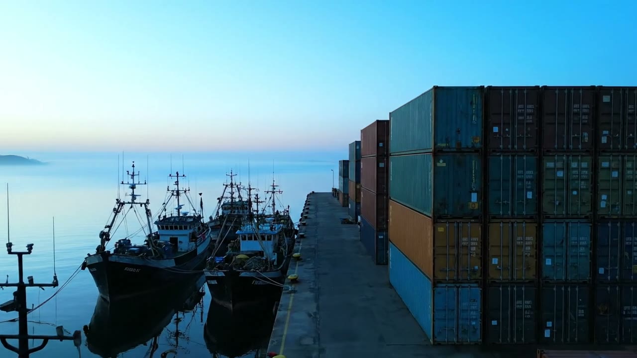 Fishing boats moored at the dock at dawn beside stacked shipping containers